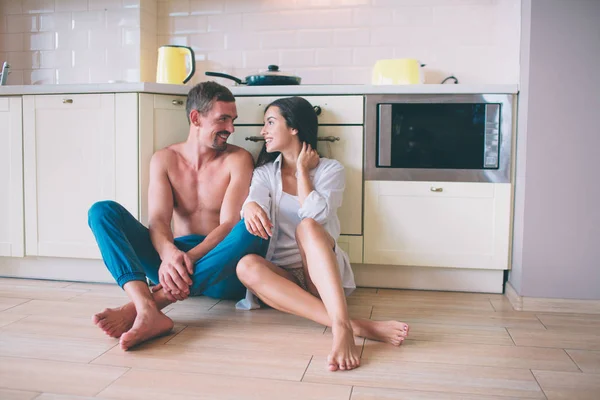 Casal encantador está sentado no chão na cozinha e olhar um para o outro. Eles mantêm as mãos cruzadas. As pessoas estão a sorrir. Parecem felizes. . — Fotografia de Stock