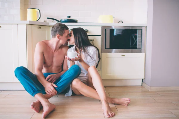 Bela foto de cara e menina sentados no chão na cozinha e beijando. Eles mantêm as pernas cruzadas. As pessoas estão inclinadas para a mobília. Ficam lindos juntos. . — Fotografia de Stock