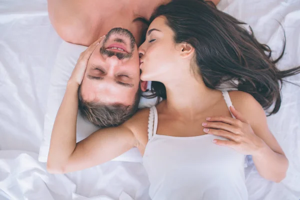 Vacker man och kvinna ljuger i vit säng. Killen håller slutna ögon och ler. Flickan är att röra vid kinden och kyssar. De är nöjda. — Stockfoto