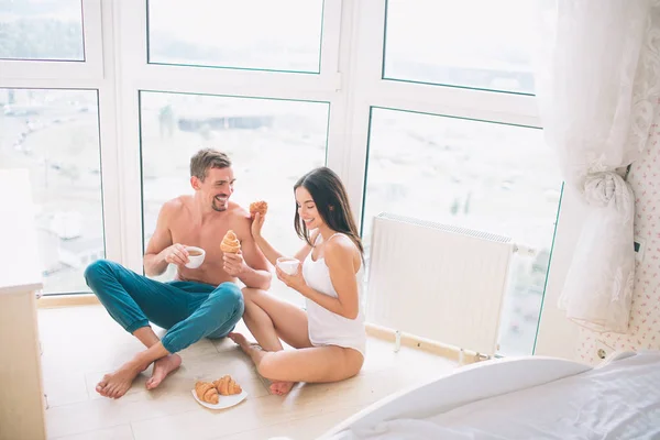 Adam ve kız pencerede oturuyor ve brekfast sahip. Croissans yemek ve beyaz bardak tutun. Kız gülümseyerek ve aşağı bakıyor. O omzuna dokunur. Biri lughing ve seyir ona. — Stok fotoğraf