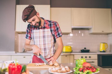 Şef pron içinde mutfak ve kesim mantarlar içerisindekiler masada bıçakla duruyor. Bu işlem ve gülümsüyor görünüyor. Olumlu biri. O yemek ve gıda hazırlama.