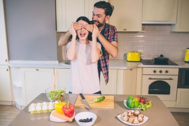 Adam kızın arkasında duruyor ve gözlerinde ellerini tutar. Heyecanlandı. Kız anlamına gelir ve elleri ayakları üzerinde duruyor. Bir sürü farklı çiğ sebze masada.