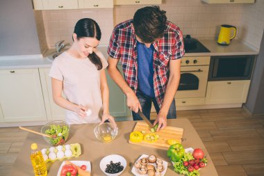 Konsantre çift masada duruyor ve birlikte çalışır. Adam yeşil salatalık parçaları keser. Kız çiğ yumurta ceket ve cam kaseye koyun. Onlar aşağı bakmak.