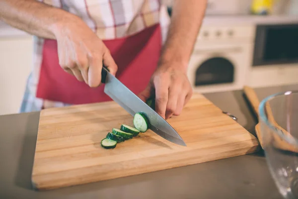 Stäng av mans händer som håller kniven och skära gurka i bitar. Han gör det på skrivbord. Det finns en glasskål på bordet. Killen bär pron. — Stockfoto