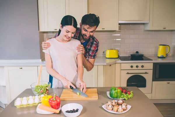 Excited and bearded guy stands behind girl and looks at food. He holds woman on shoulders. Girl looks down as well. She cuts some cheese on desk. Girl is cooking. — Stock Photo, Image