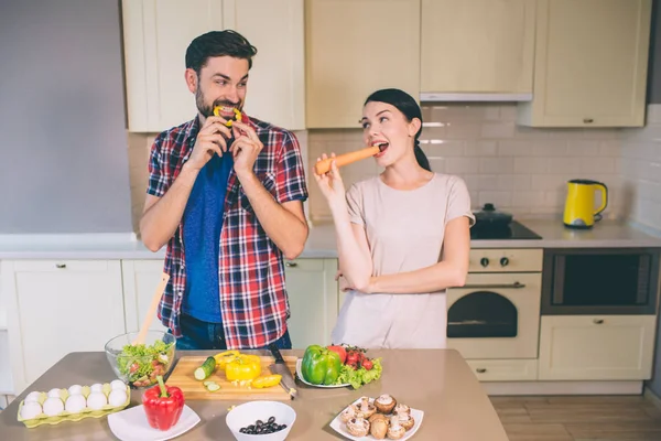 Dark-haired members of one couple stands and looks at each other. They are biting piece of yellow pepper and orange carrot. People look funny. They are enjoying company of one another. — Stock Photo, Image