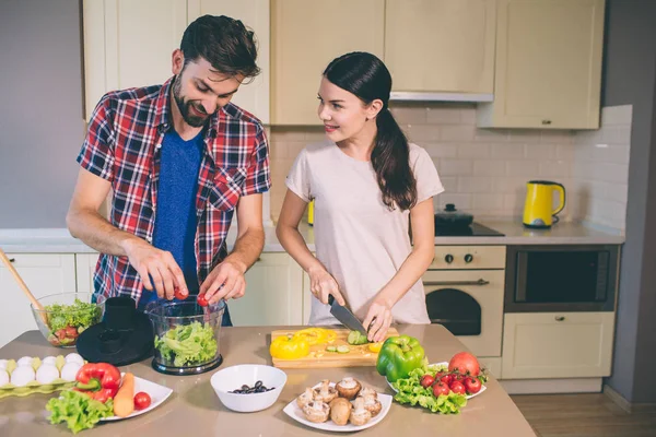 Ωραίο ζευγάρι δουλεύει μαζί στην κουζίνα. Αυτή περικοπές μανιτάρια και κοιτάξτε ο τύπος. Είναι juicying ντομάτες στο πλαστικό μπολ και ματιά. Είναι συγκεντρωμένοι. — Φωτογραφία Αρχείου