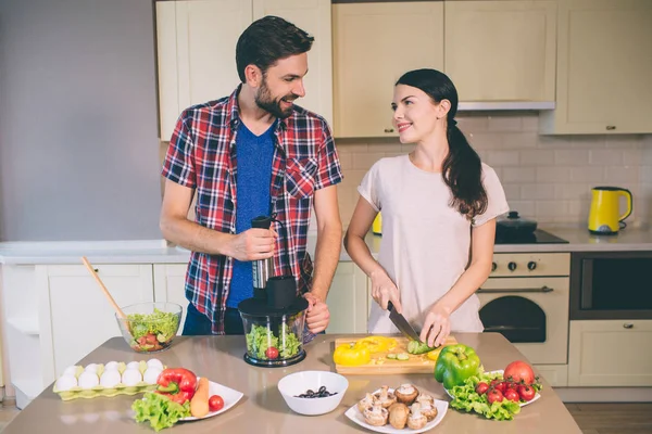 Delightful couple works together. Girl is cutting cucumber into pieces and looks at guy. He holds ingredients for salad in blender. Man looks at woman. They smile to each other. — Stock Photo, Image