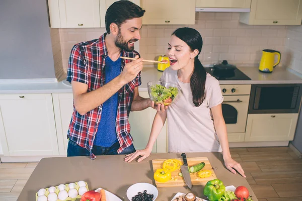 Cute picture of man standing at table and holding bowl with salad. He is stretching hand with wooden spoon with tomato on it. Girl opens her mouth. She wants to taste it. — Stock Photo, Image