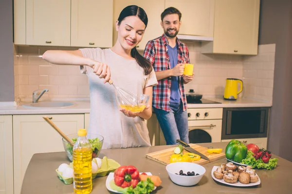 Beautiful woman stands and smashes eggs in glass bowl. She looks at it and smile. Guy stands behind her. He leans at kitchen furnoture and smiles. Man holds yellow cup in hands. — Stock Photo, Image