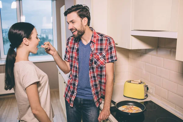 Lovely pictue of man standing in front of woman and holding wooden spoon with piece of omlet. She looks at it and keeps mouth opened. Girl is going to taste omlet. Guy is smiling. He is excited. — Stock Photo, Image