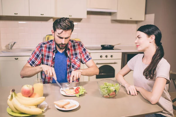 Hungry man sits and table and cuts meal in his plate with fork and knife. Guy is concentrated. Girl sits besides him and looks at man. She is happy. — Stock Photo, Image