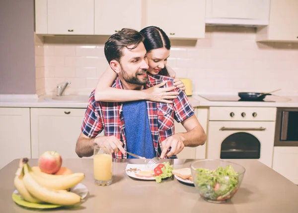 Careful girl stands behind guy and embrace him. He looks to the right and smile a bit. He cuts meal on his plate. Man is eating. — Stock Photo, Image
