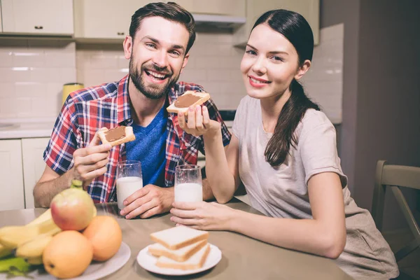 Positive picture of guy and girl sits together at tble and smiling. They look on camera. People hold toast with chocolate paste and glasses of milk. They are gorgeous. — Stock Photo, Image