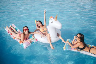 Seksi esmer yüzme havuzunda yüzen üzerinde. Onlar birlikte tüyler ürpertici. Orta noktaları yukarı ve gülümseyen kadın. Diğer modeller kamerada poz olan. Onlar mutlu.