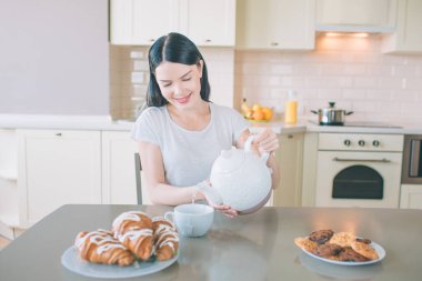 Pozitif kadın mutfakta masada oturur. O su beyaz fincan içine gülümsüyor ve poures. Tabak kurabiye nad croissans masada ile vardır. Mutlu.