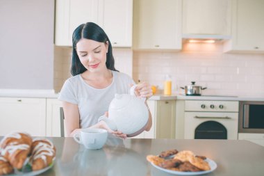 Sakin ve huzurlu esmer masada oturur ve aşağı görünüyor. O beyaz su ısıtıcısı sahiptir. Kadın poures ondan bardağa su. Konsantre görünüyor.