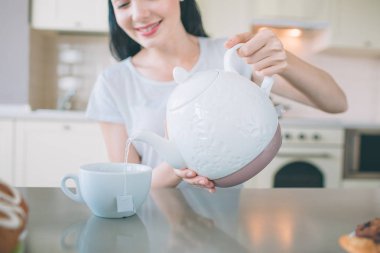 Esmer kesme görünümü masada oturur. O beyaz su ısıtıcısı tutar ve bardağa su dökülen. Kız çay hazırlanıyor. O sitd mutfak.