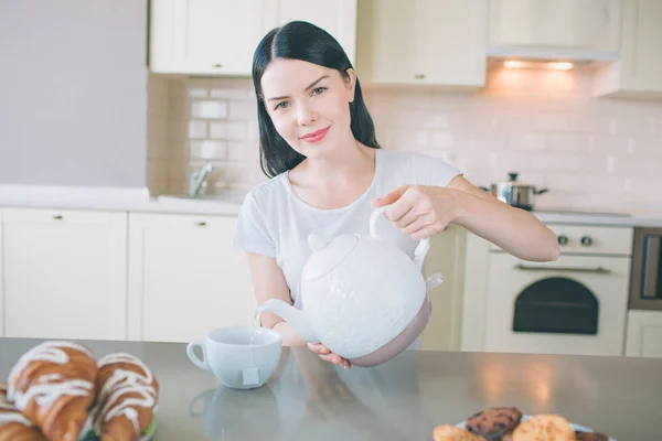 Mulher jovem positiva senta-se à mesa na cozinha e olha para a câmera. Ela segura a chaleira branca e derrama água no copo. Mulher posa. Ela está bonita. . — Fotografia de Stock