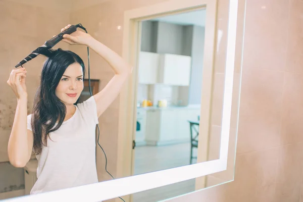 Brunette se tient au miroir et en utilisant des cheveux bouclés. Elle est dans la salle de bain. Femme tient Curler avec les deux mains. Elle fait de la coiffure . — Photo