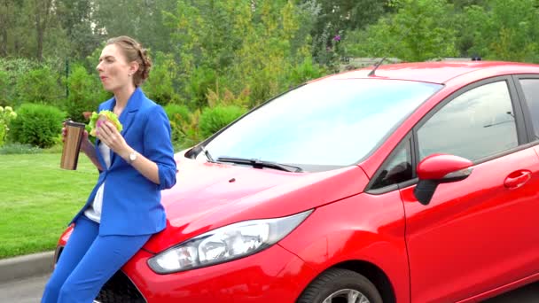 Frau im blauen Anzug sitzt auf Motorhaube eines roten Autos und isst zu Mittag. Mädchen isst Sandwich und trinkt aus Thermoskanne. Sie ist ruhig und gelassen. Mädchen blickt geradeaus. — Stockvideo