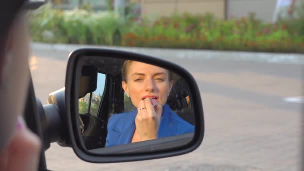 Aantrekkelijk meisje zit in de auto en maakt gebruik van lippenstift. Ze doet make-up voor haar lippen. Meisje kijkt in de spiegel. — Stockvideo