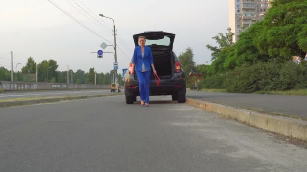 Mulher de fato azul caminha para a câmara. Ela tirou o triângulo vermelho do anúncio do porta-malas. Mulher coloca-o no chão e mantém a mão de lado. Ela quer parar o carro. . — Vídeo de Stock