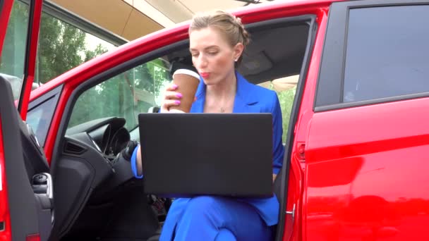 Дівчина сидить у машині з відкритими дверима. Вона п'є каву з чашки і дивиться на екран ноутбука. Тоді жінка знову п'є і продовжує працювати . — стокове відео