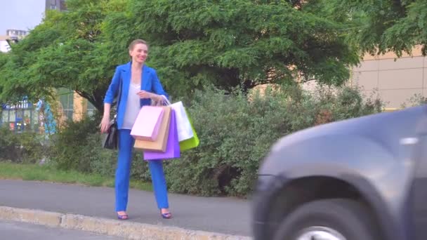 Mutlu bir kadın duruyor ve alışveriş torbaları tutar. O gülümsüyor. El ile kız dalgalar. O araba durduktan sonra. İçinde oturuyor. Daha ileri gitmek o arabayı başladıktan sonra. — Stok video