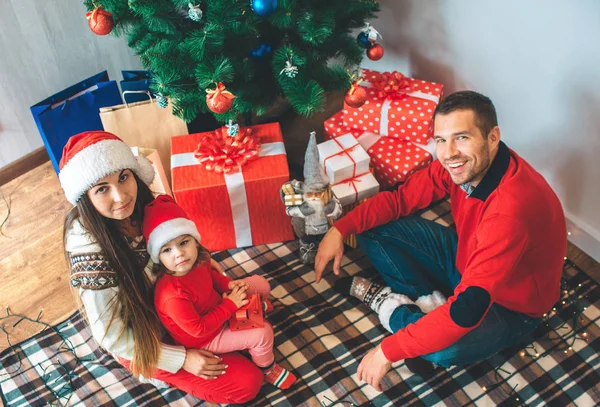 Joyeux Noël et bonne année. Photo festive d'adultes et de filles assis sur une couverture et levez les yeux sur la caméra. Un homme sourit. Il s'assoit de femme en fille. Ils sont près des boîtes rouges avec des cadeaux et des arbres . — Photo