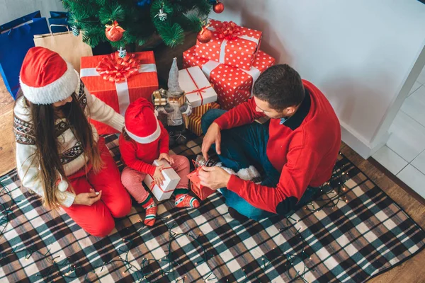 Joyeux Noël et bonne année. Le gamin est assis entre parents et cadeau ouvert. Elle est concentrée. Il tient une lanterne rouge et regarde vers le bas. Mère s'assoit et regarde l'enfant. Elle porte un chapeau de Noël . — Photo