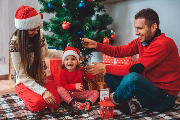 Frohe Weihnachten und ein gutes neues Jahr. glückliches kleines Mädchen sitzt zwischen Eltern und lacht. Mutter blickt nach unten. Sie trägt Hut. Kerl im roten Pullover lächelt und hält Weihnachtslicht auf Mädchenkopf. — Stockfoto