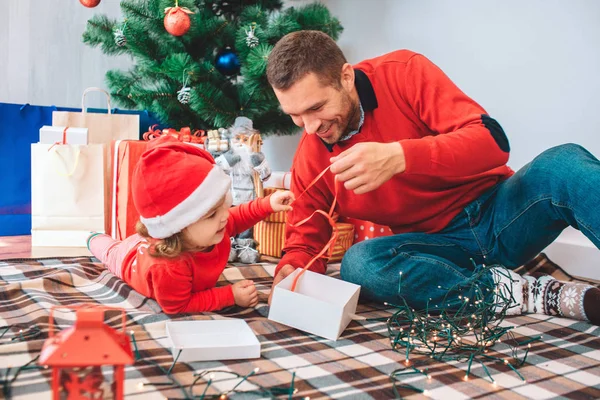 Joyeux Noël et bonne année. Le gamin et son parent sont assis ensemble sur une couverture et tiennent un ruban rouge. Ils le regardent. Le ruban a été montré de la boîte blanche. Un homme sourit. Fille est intéressé par la boîte — Photo