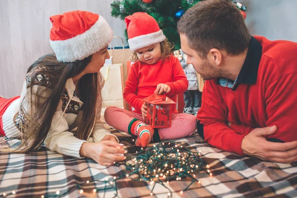 Frohe Weihnachten und ein gutes neues Jahr. Konzentriertes Kind sitzt zwischen Eltern. Sie hält die rote Laterne und spielt damit. Frau und Mann betrachten das Kind. Sie sind vorsichtig. Mutter und Kind tragen Weihnachtsmützen. — Stockfoto
