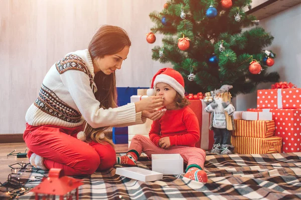 Joyeux Noël et bonne année. Belle image de la mère et de l'enfant assis ensemble. La femme nourrit la petite fille. Le gamin mâche. Les filles sont à l'arbre de Noël avec de nombreuses boîtes rouges . — Photo