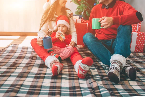 Joyeux Noël et bonne année. Couper la vue de la famille assise ensemble sur une couverture. Ils portent des vêtements colorés. Des hommes et des femmes tiennent des tasses. Petite fille assise sur les genoux des mères. Elle boit du jus. Enfant — Photo