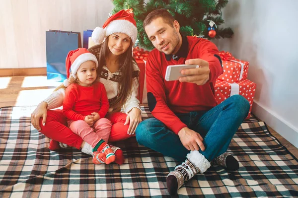 Joyeux Noël et bonne année. Le jeune homme est assis à côté de la femme et de l'enfant. Il tient son téléphone et prend du selfie. Femme et enfant s'y attardent et posent. La famille porte des vêtements et des chapeaux de Noël . — Photo