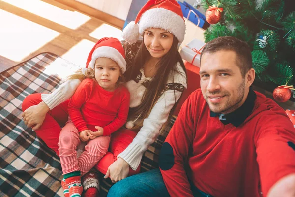 Frohe Weihnachten und ein gutes neues Jahr. Bild einer netten Familie. junger Mann hält Kamera und macht Selfie. Sie alle posieren. Kind sieht ernst aus. Sie sitzen am Weihnachtsbaum auf einer Decke. — Stockfoto