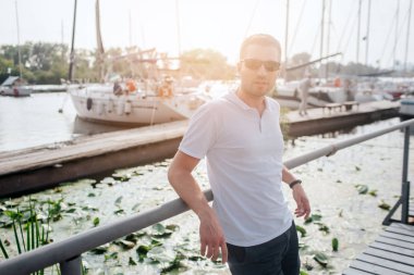 Güzel ve yakışıklı adam iskelede durmak ve korkuluk yalın. Kameraya bakıyor. Kendine güvenen biri. Beyaz gömlek ve siyah şort giyiyor.