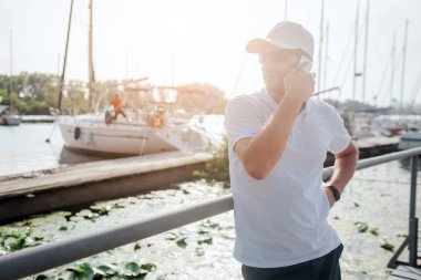 İskele üzerinde genç adam duruyor meşgul ve sola bakın. Telefonla konuşuyor. Yatlar arkasında vardır. Güneş batıyor. Adam bekliyor.