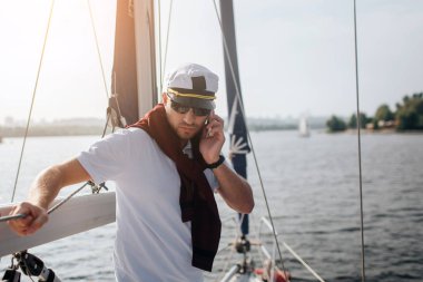Ciddi ve yoğun yachtsman standları ve parmaklıklara yaslan. Elinde telefon tutun ve aşağı bak. Adam çalışıyor. ITIS dışında günbatımı.