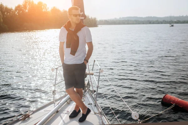 时尚的年轻水手站在船头的游艇。他摆出姿势, 往一旁看。那家伙戴墨镜他已经准备好去旅行了。太阳下山了. — 图库照片