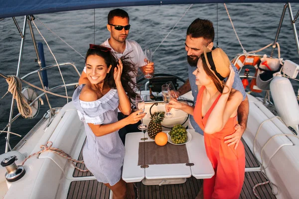 Två par på yacht har part. De innehar glases av champaigne. Kvinnor ler. De ser glada. Män står med dem och omfamna sina flickvänner. — Stockfoto