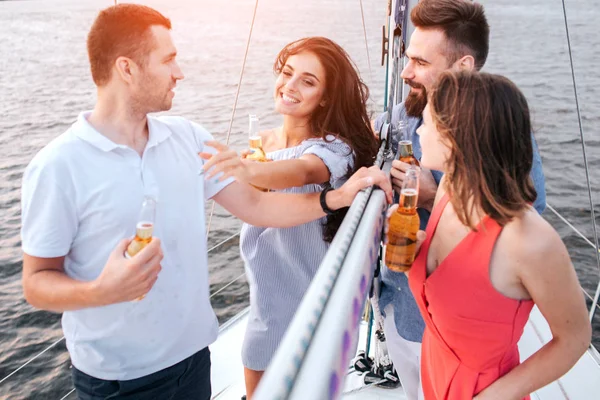 Beautiful picture of brunette touches man and smiles. Another couple standing in front of them. They have bottles of beer. People relax and chill.