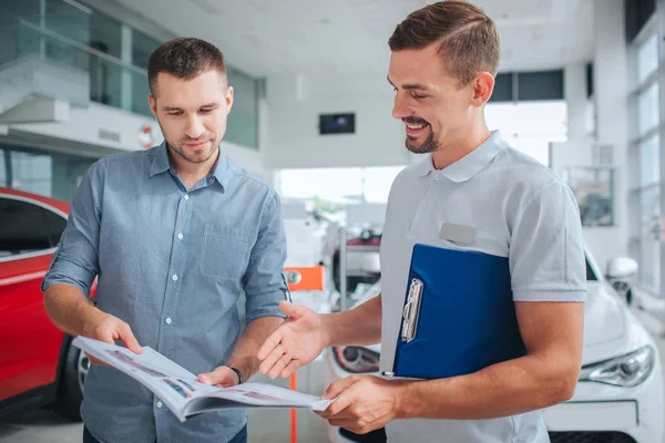 Resmi içinde duran iki adam. Onlar günlük tutun ve ona bak. İnsanlar gülümse. Kırmızı araba duruyorlar. Günlük ve gülümseme beyaz gömlek puan erkekte.