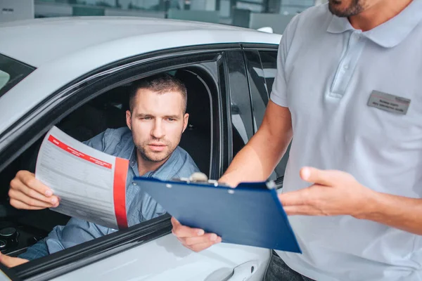 Venditore sta in piedi a macchina bianca e tiene tablet in mano. L'ha indicato lui. Un altro uomo si siede in macchina e detiene carta di assicurazione. È serio e concentrato. Hanno una conversazione. . — Foto Stock