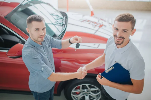 Acquirente e commerciante in piedi e stringersi la mano. Venditore detiene la chiave dalla macchina. Guardateli entrambi davanti alla telecamera. Sono vicino alla macchina rossa. Entrambi i giovani sono felici . — Foto Stock