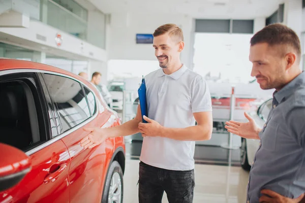 Araba satıcı ve müşteri güzel kırmızı araba ile stand ve bakmak. Onlar gülümseme. Danışman plastik mavi tablet tutar. Müşteri noktası eliyle. — Stok fotoğraf