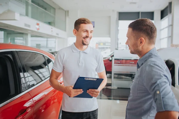 Εικόνα του πελάτη και ο πωλητής ενωμένοι μπροστά από το όμορφο κόκκινο αυτοκίνητο. Φαίνονται σε κάθε άλλο και χαμόγελο. Πωλητής κρατά πλαστική tablet. Είναι στο σαλόνι. — Φωτογραφία Αρχείου