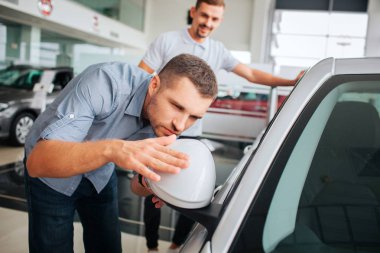 Müşteri beyaz araba üzerinde eğilir ve ona dokunursa detaylara özenli. Genç adam aynaya bak. O ciddi ve konsantre. Satıcı onu ve gülümsüyor duruyor. Çok olumlu olduğunu.